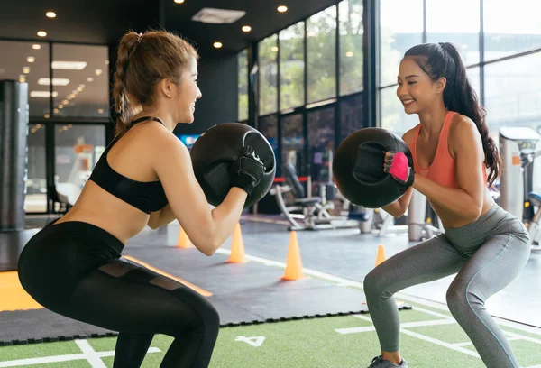 Happy young female athletic people performing squat exercises with friend and holding a medicine ball at fitness gym. Group of two confident women with healthy lifestyle working out together.