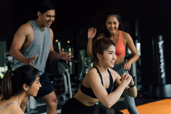 Group of people cheering on their Asian female friend doing squats with a weight plate in fitness gym. Working out together as a teamwork.