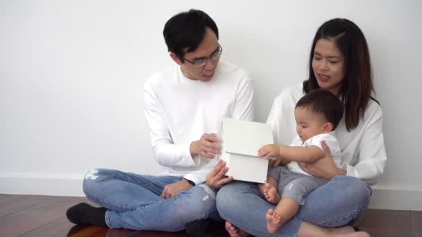 Alegre mãe étnica asiática e pai sorrindo e mostrando casa de brinquedo para bebê bonito enquanto sentado cruzou as pernas no chão perto da parede branca em casa — Vídeo de Stock