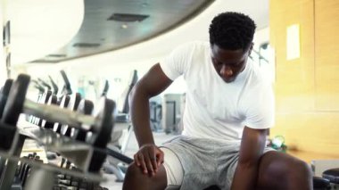Genç Afrikalı Amerikalı bir adam spor salonundaki rafa yakın bir yerde oturuyor ve dambılı kaldırıyor. Vücut geliştirme merkezinde biceps kıvıran erkek ağırlık eğitmeni.