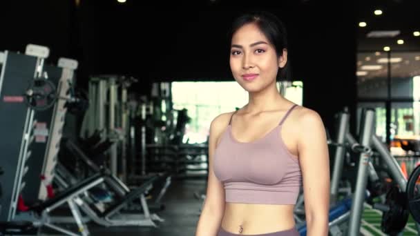 Sonriente y decidida joven atleta asiática mirando a la cámara mientras pone las manos en las caderas y de pie en el gimnasio gimnasio — Vídeo de stock