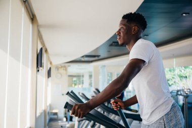 Modern spor salonunda spor giyinmiş, spor yaparken eliptik makineye basamaklar yapan Afro-Amerikan bir adamın yan görüntüsü.