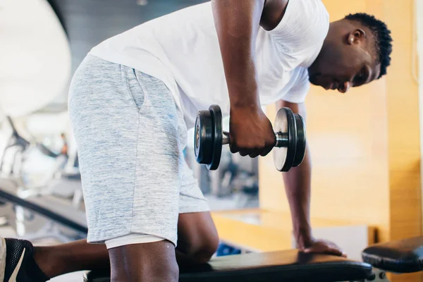 Вид сбоку на афроамериканца в спортивной одежде, опирающегося на скамейку и делающего упражнения с гантелями во время тренировки в спортзале — стоковое фото