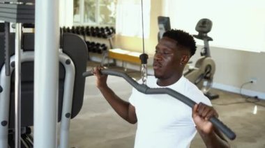 Spor giyim sektöründeki güçlü Afro-Amerikalı adam spor salonunda antrenman sırasında egzersiz makinesinde otururken çubukla kablo çekiyor.