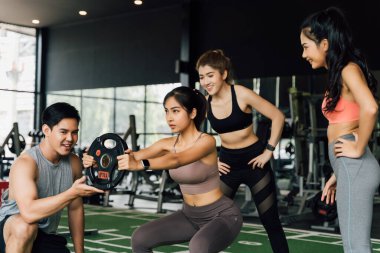 Bir grup insan, spor salonunda ağırlık plakasıyla çömelme hareketi yapan Asyalı bayan arkadaşına tezahürat yapıyor. Takım çalışması olarak çalışıyoruz..