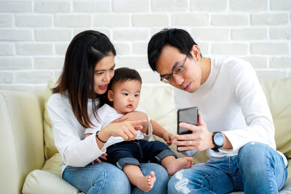 Alegre joven familia asiática con hijo viendo vídeo en el teléfono móvil y sentado en sofá sofá en casa —  Fotos de Stock
