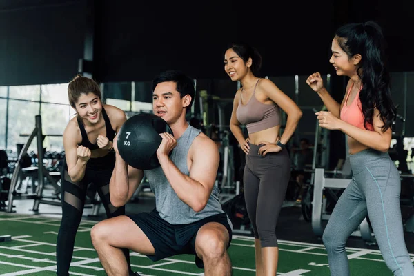 Een groep mensen juichen hun Aziatische Chinese mannelijke vriend toe die hurkt met een medicijnbal in de fitnessruimte. Samenwerken als een teamwork. — Stockfoto