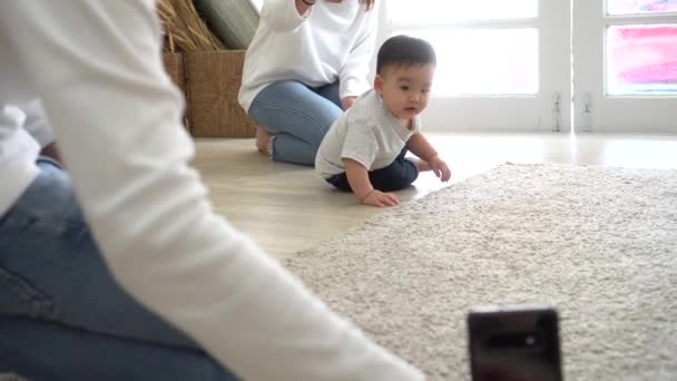 Eltern filmen mit Smartphone süßes asiatisches Baby, das zu Hause auf weichem weißen Teppich krabbelt — Stockvideo