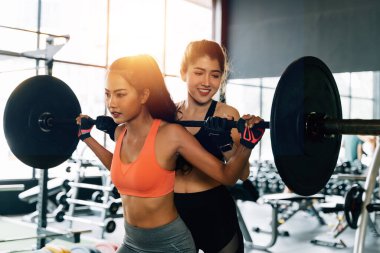 Asyalı kişisel antrenör vücut geliştiren bir kadına spor salonunda halterle egzersiz yapması için koçluk yapıyor..