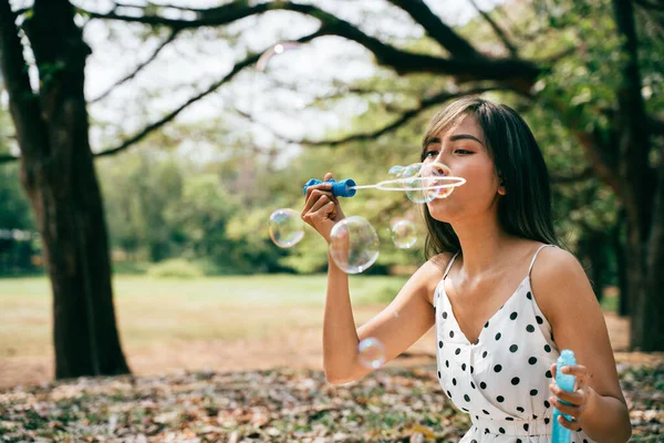 Jovem do Sudeste Asiático étnico 20 anos mulher soprando bolhas de ar no parque. Menina feliz usa vestido de verão com árvores no fundo. — Fotografia de Stock