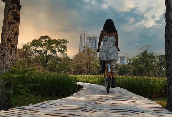 Siluet wanita muda mengendarai sepeda di jalan pedesaan dengan bangunan perkotaan di latar belakang pengendara motor perempuan yang sehat mengenakan gaun musim panas saat matahari terbenam — Stok Foto