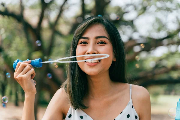 Jovem do Sudeste Asiático étnico 20 anos mulher soprando bolhas de ar no parque. Menina feliz usa vestido de verão com árvores no fundo. — Fotografia de Stock