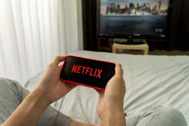 BANGKOK, THAILAND - 27 Mart 2020: yatakta oturan adam Netflix logosunu akıllı telefon ekranında tutuyor. Netflix internet üzerinden yayın yapan bir eğlence şirketidir.