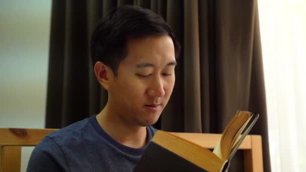 Porträtt av fridfulla 30-talets asiatiska man i vardagskläder tillbringar fritiden med att läsa en bok hemma på sängen. Rekreation och fritid — Stockvideo