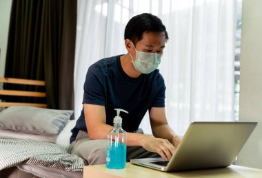 Asyalı adam yatak odasında koruyucu maske takarak çalışıyor. Evde koruyucu maske takarak koronavirüs için karantina. Ellerini temizlemek için dezenfektan jeli kullanıyor..