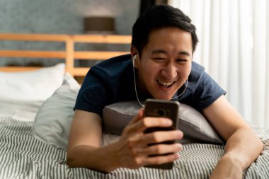 30 'lu yaşların mutlu Asyalı adam portresi. Günlük kıyafetleri içinde. Evde akıllı telefonuyla görüntülü arama yapıyor. Telefon ekranındaki insanlara el sallıyor..
