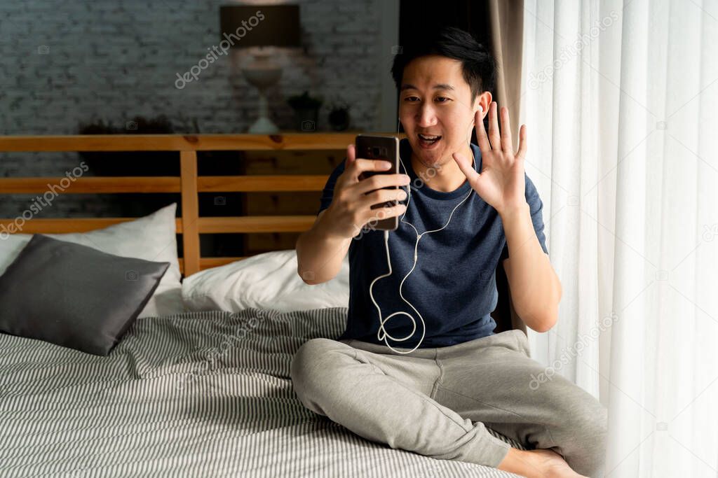 Portrait of happy 30s aged Asian man in casual clothing making facetime video calling with smartphone at home. Hes waving at people on phone screen.