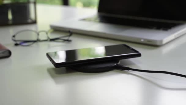 Chytrý telefon je nabíjen na bezdrátové nabíječce spolu s notebookem, brýlemi a notebookem na stole — Stock video