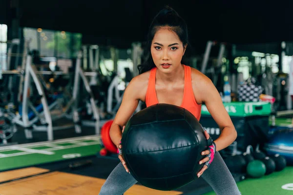 Entschlossene und ernsthaft aktive asiatische Sportlerin hockt beim Training im Fitnessclub mit Medizinball — Stockfoto