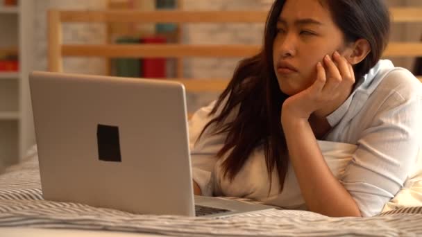 Retrato de la joven asiática aburrida acostada en la cama mientras trabajaba desde casa en el dormitorio. freelancer femenina que trabaja en la computadora en línea. Concepto de expresión frustrado y acentuado — Vídeos de Stock