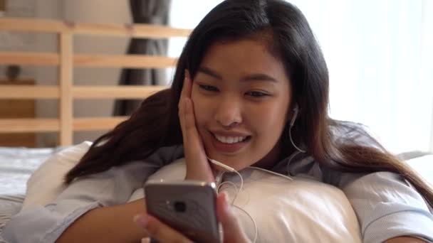 Retrato de la joven asiática feliz con ropa casual acostada en la cama mientras hace una videollamada con teléfono inteligente y saludando en el dormitorio en casa. Concepto de tecnología de videoconferencia — Vídeo de stock