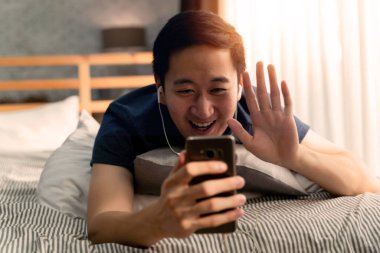 30 'lu yaşların mutlu Asyalı adam portresi. Günlük kıyafetleri içinde. Evde akıllı telefonuyla görüntülü arama yapıyor. Telefon ekranındaki insanlara el sallıyor..