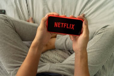 BANGKOK, THAILAND - 27 Mart 2020: yatakta oturan adam Netflix logosunu akıllı telefon ekranında tutuyor. Netflix internet üzerinden yayın yapan bir eğlence şirketidir.
