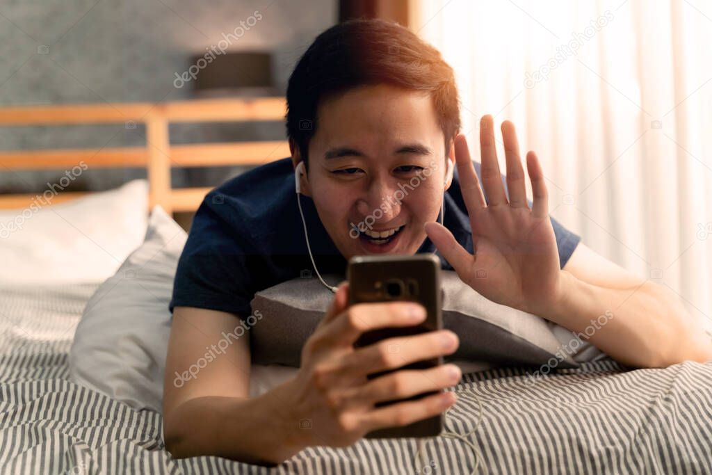 Portrait of happy 30s aged Asian man in casual clothing making facetime video calling with smartphone at home. Hes waving at people on phone screen.