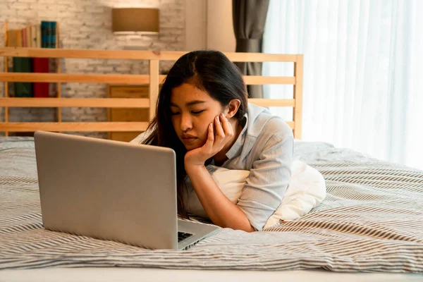 Портрет скучающей молодой азиатки, лежащей на кровати, работающей из дома в спальне. Женщина-фрилансер работает на компьютере онлайн. Концепция выражений разочарования и стресса — стоковое фото