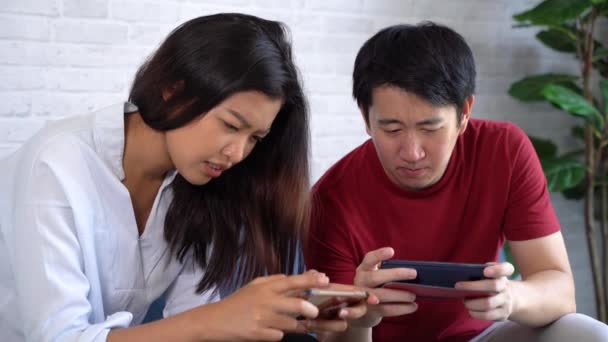 Jóvenes amigos y amigas jugando en el teléfono móvil en casa. Asiático hombre y mujer usando smartphone para jugar videojuego juntos — Vídeos de Stock
