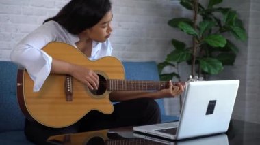 Genç Asyalı kadın dizüstü bilgisayar monitöründen gitar çalmayı öğreniyor. Kadın gitarist çevrimiçi özel ders izliyor.