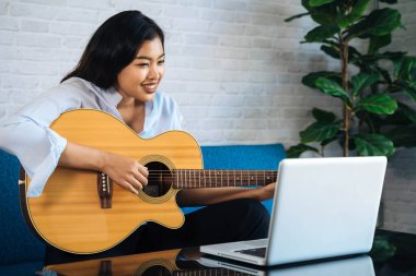 Genç Asyalı kadın dizüstü bilgisayar monitöründen gitar çalmayı öğreniyor. Kadın gitarist çevrimiçi özel ders izliyor.