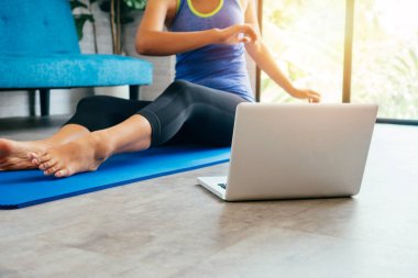 20 'li yaşlarda, spor giyimli genç bir Asyalı kadın bilgisayar laptopunda yoga eğitimi alırken stering egzersizleri yapıyor. Arka planda kanepe ile oturma odasında egzersiz yapan sağlıklı bir kız.