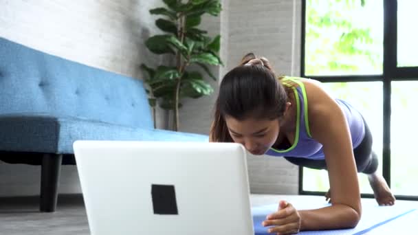 20-talet ung asiatisk kvinna i sportkläder gör planka poser medan du tittar på fitness träningsklass på datorn bärbar dator på nätet. Friska flicka utövar och lär i vardagsrummet. Internetutbildningskoncept — Stockvideo