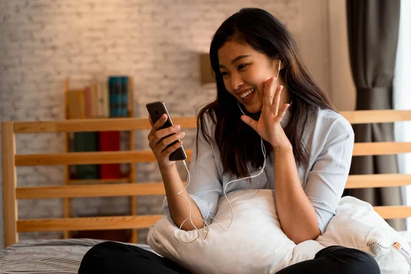 평범 한 옷을 입고 침대에 앉아 있는 행복 한 아시아 소녀의 모습 집의 침실에서 스마트폰으로 비디오 전화를 하는 모습. 비디오 콘퍼런스 기술 개념 — 스톡 사진