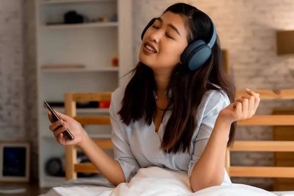 Jovem mulher asiática feliz em fones de ouvido cantando e ouvindo música enquanto está sentado na cama e segurando o telefone celular em casa — Fotografia de Stock