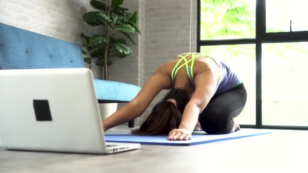 20-летняя молодая азиатка в спортивной одежде делает упражнения на растяжку во время просмотра занятий йогой на компьютерном ноутбуке онлайн. Здоровые девушки упражнения в гостиной с диваном на заднем плане — стоковое видео
