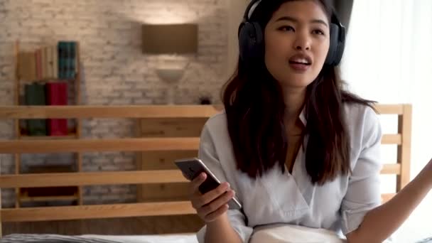 Ευτυχισμένη νεαρή Ασιάτισσα με ακουστικά που τραγουδάει και ακούει μουσική ενώ κάθεται στο κρεβάτι και κρατάει το κινητό τηλέφωνο στο σπίτι — Αρχείο Βίντεο