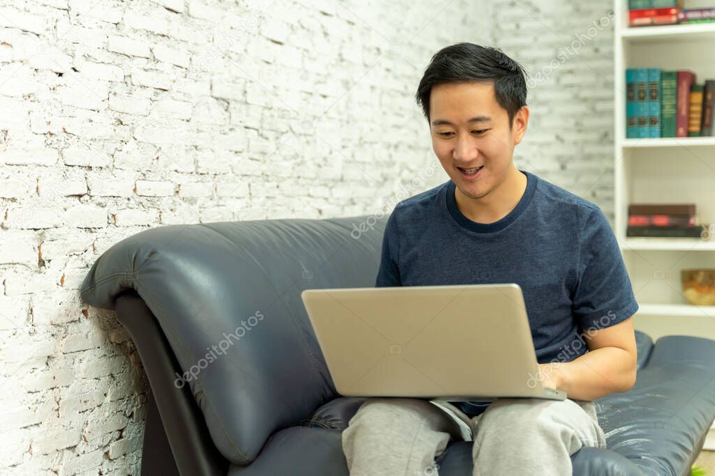 Young smiling Asian man in casual clothing on sofa working on laptop computer. Male freelancer working from home on notebook.
