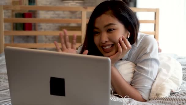 Retrato de la joven asiática feliz en ropa casual acostado en la cama mientras que hace una videollamada con computadora portátil en el dormitorio en casa. Concepto de tecnología de videoconferencia — Vídeo de stock