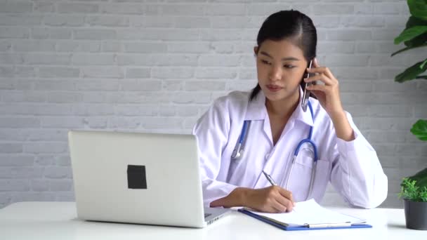 Doctora joven enfocada hablando por teléfono móvil y escribiendo notas en el hospital. Mujer asiática concentrada usando un portátil en el consultorio médico — Vídeos de Stock
