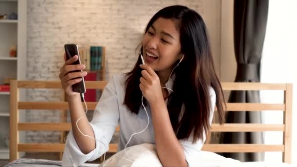 Portrait of happy young Asian girl in casual clothing lying down on bed while making a video call with smartphone in bedroom at home. Video conferencing technology concept — Stock Video