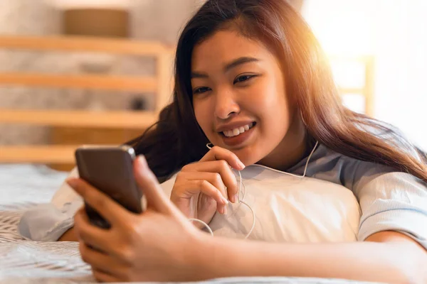 Портрет счастливой молодой азиатки в повседневной одежде, лежащей на кровати во время видеозвонка со смартфона и машущей в спальне дома. Концепция технологии видеоконференций — стоковое фото