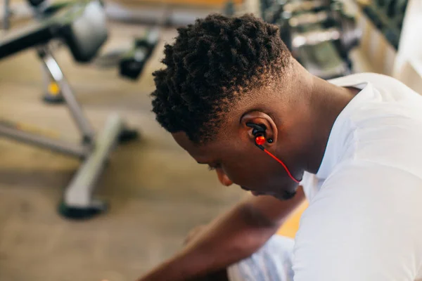 Vista lateral do homem afro-americano cansado em sportswear ouvir música em fones de ouvido sem fio enquanto sentado no banco e descansando durante o treinamento em ginásio — Fotografia de Stock