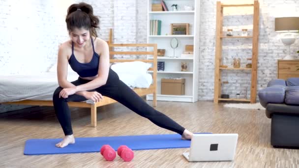 20-talet ung asiatisk kvinna i sportkläder gör stretching övningar medan du tittar på fitness träningsklass på datorn bärbar dator på nätet. Friska flicka utövar i vardagsrummet med soffa soffa och säng — Stockvideo