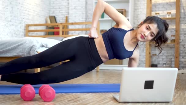 20s mladá asijská žena ve sportovním oblečení dělá strečink při sledování fitness trénink na notebooku online. Zdravá dívka cvičení v obývacím pokoji s pohovkou v pozadí — Stock video