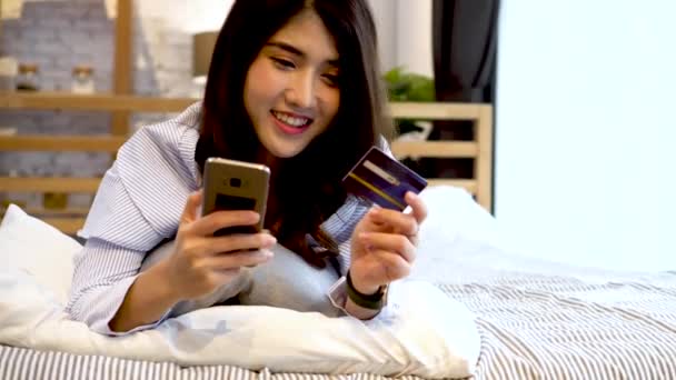 Retrato de feliz 20 anos mulher asiática em roupas casuais segurando um cartão de crédito e telefone celular na cama em casa. Finanças, comércio eletrônico, banco de dinheiro conceito on-line — Vídeo de Stock