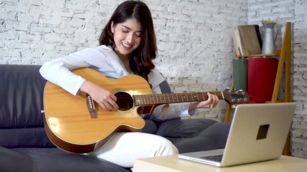 Mujer asiática joven practicando y aprendiendo a tocar la guitarra en el monitor del ordenador portátil. Guitarrista femenina viendo tutorial en línea — Vídeo de stock