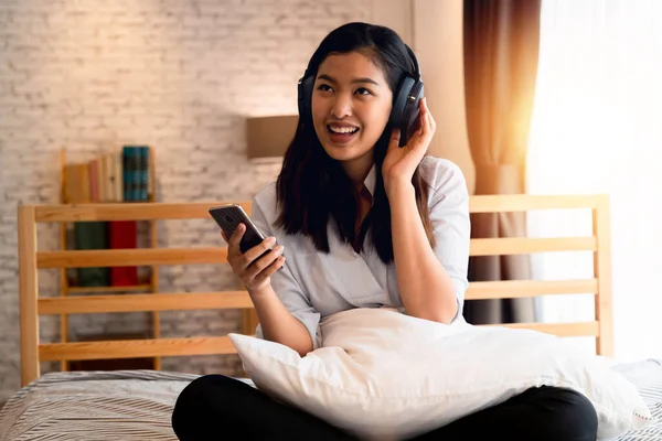 Jovem mulher asiática feliz em fones de ouvido cantando e ouvindo música enquanto está sentado na cama e segurando o telefone celular em casa — Fotografia de Stock