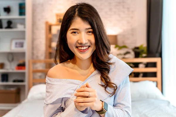 自宅のベッドルームでビデオ通話をしながら、ベッドの上に座って笑顔のアジアの少女の肖像画。ビデオ会議技術とワイヤレス通信の概念-視点のヘッドショット — ストック写真